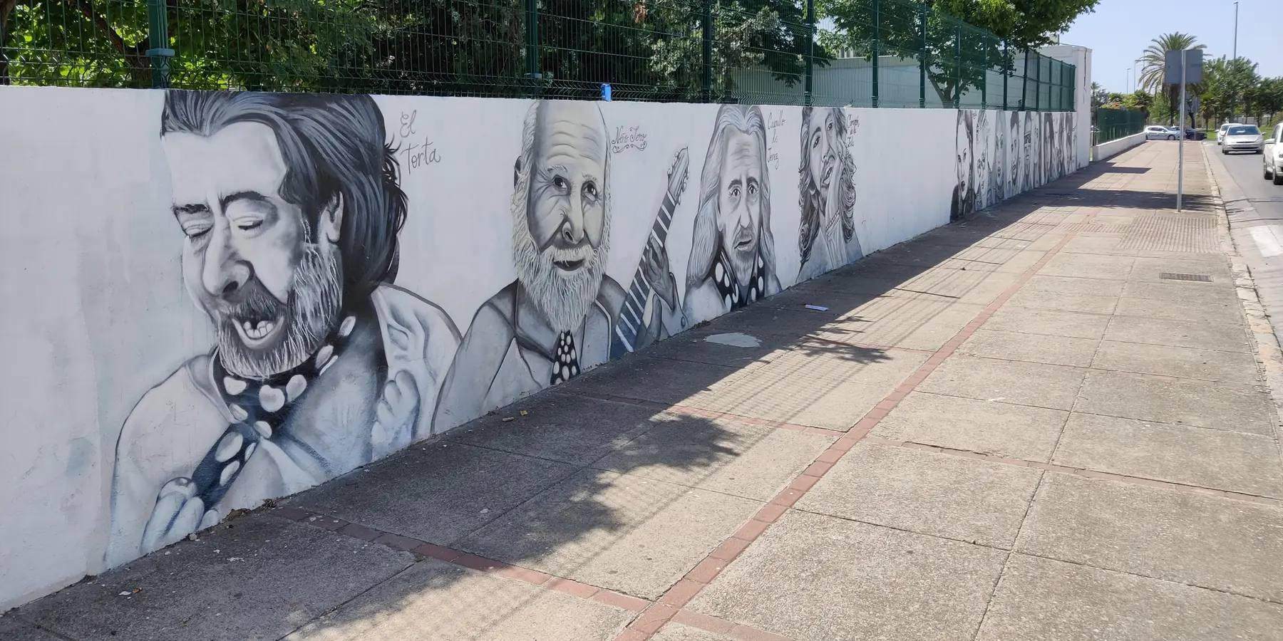 Un lungo muro imbiancato a calce di una scuola a Jerez, dipinto con ritratti grigi di cantanti flamenco e un chitarrista, visto lungo un marciapiede sotto gli alberi