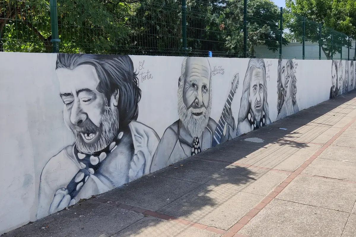 Ένας μακρύς ασβεστωμένος τοίχος σχολείου στη Jerez, ζωγραφισμένος με γκρίζα πορτρέτα τραγουδιστών και κιθαριστών του flamenco, δίπλα σε ένα πεζοδρόμιο
