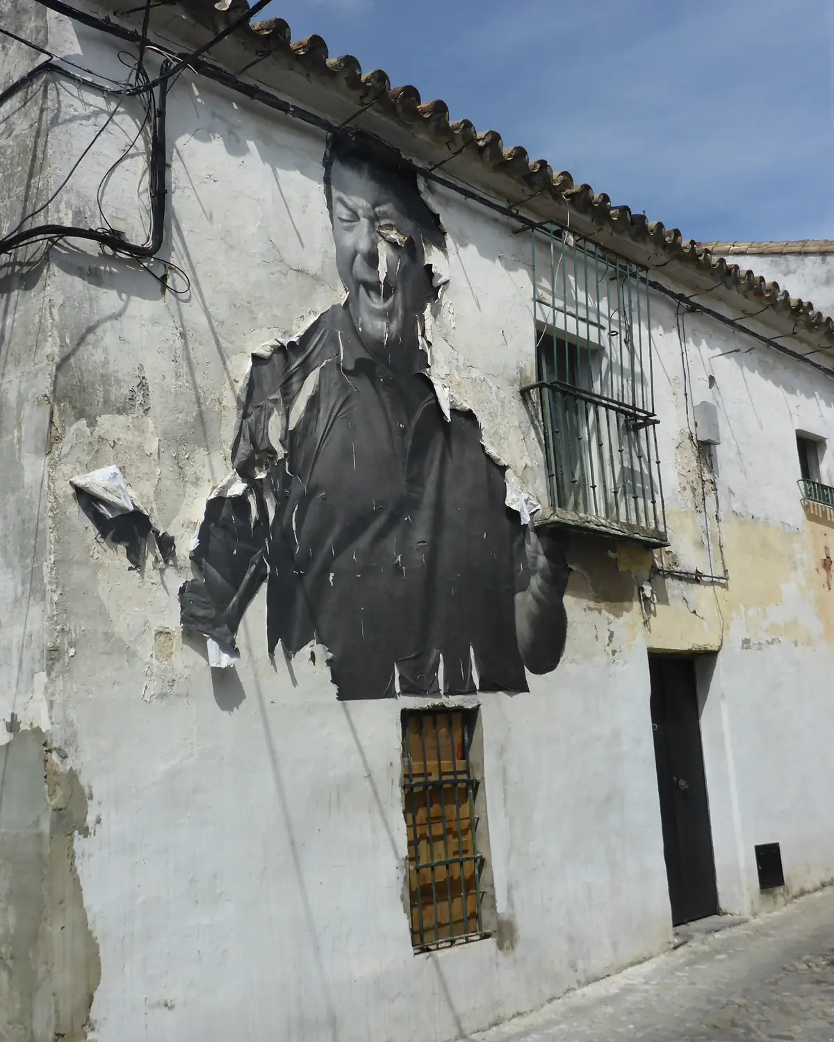 Un grande ritratto in bianco e nero di un cantante flamenco che ride, affisso sul muro scrostato e imbiancato a calce di una casa di Jerez, con una finestra con inferriata accanto